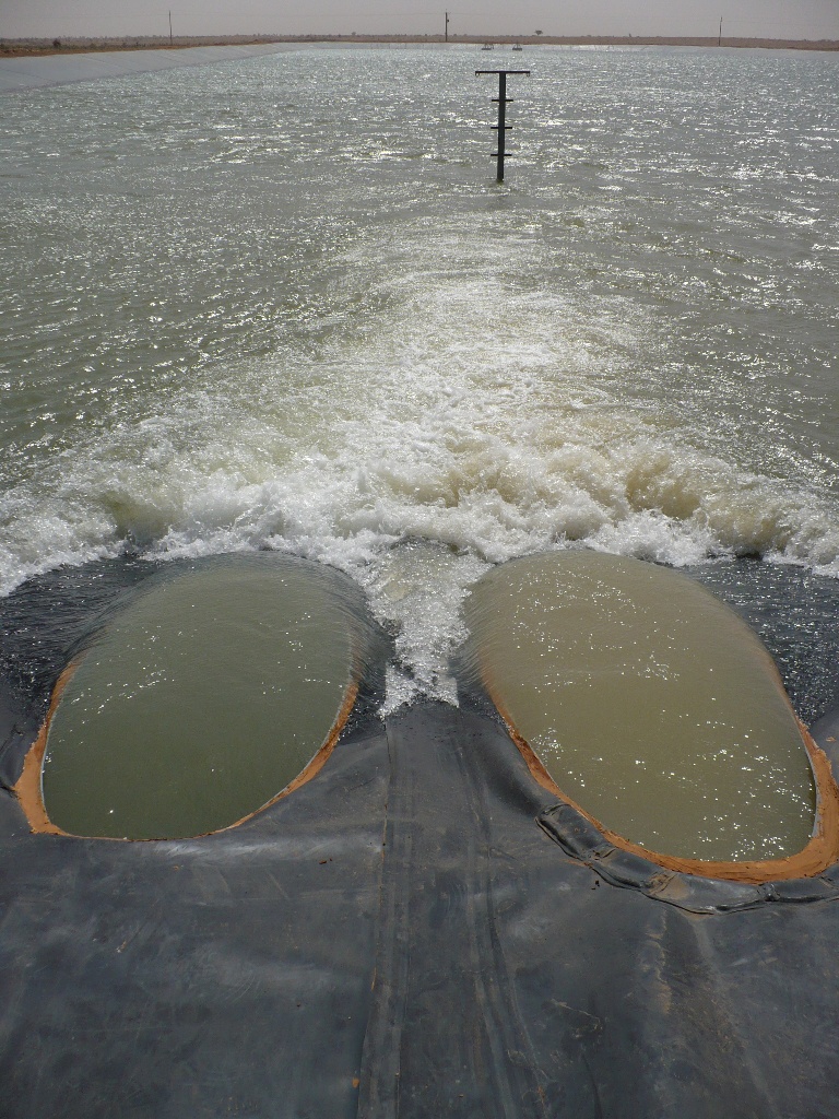 Two large pipe outlets discharging water into a lined reservoir with turbulent whitewater and a gauge post, grey and blue tones