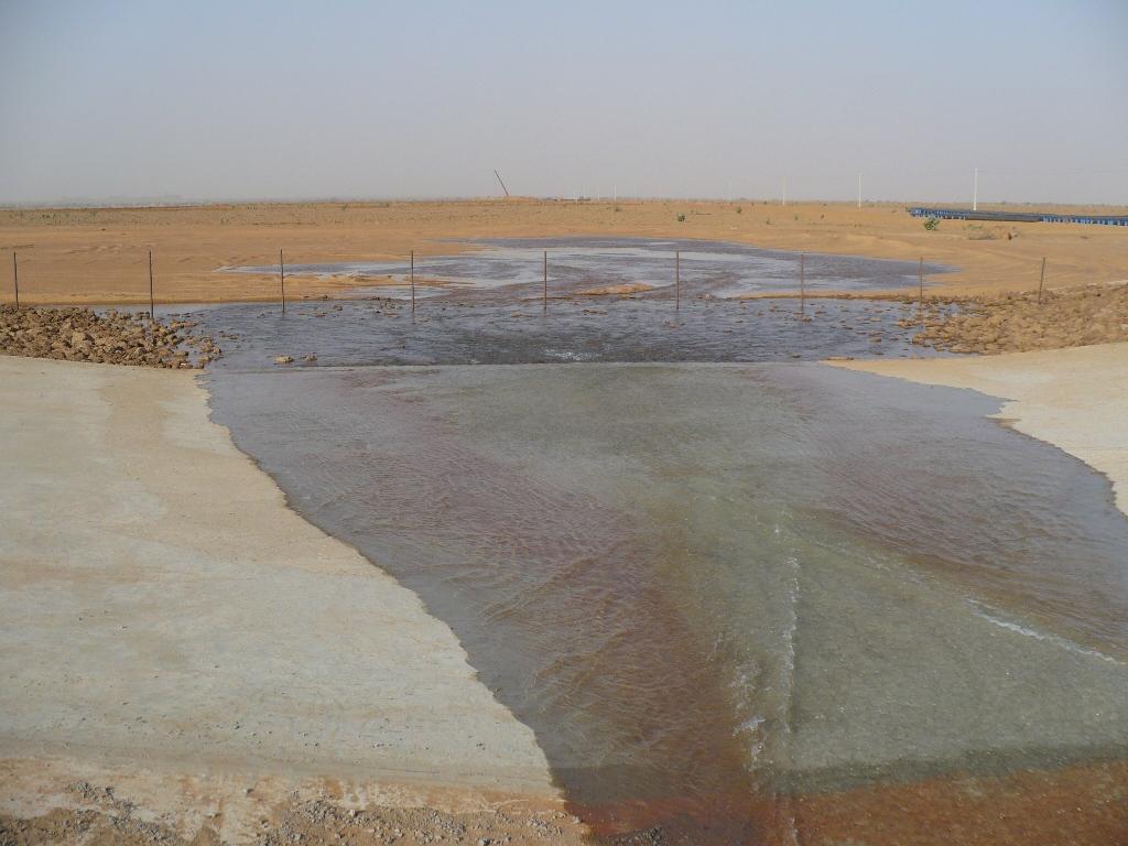 Shallow reddish-brown water pooling on a concrete surface in an open desert area with fencing and distant structures, muted brown tones